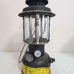 Coleman U.S. Army SMP 1984 pressure lantern with dark military-style fount, black ventilator, clear globe, wire frame and yellow instruction label. Shows paint wear, oxidation and general age-related 
