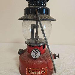 Kamplite Model LRL-21A pressure lantern with red fount, black porcelain ventilator, clear globe and wire bail handle. Shows paint wear, oxidation and age-related use.